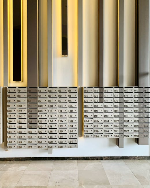 A row of silver parcel lockers with numbered compartments, mounted on a white wall beneath vertical gold and black decorative panels, in a well-lit indoor setting. The lockers are used for secure flow