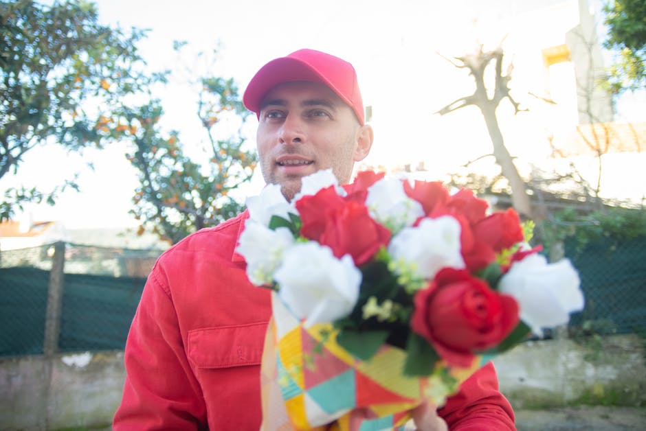 A male flower delivery courier dressed in a red uniform and cap, holding a vibrant bouquet of fresh roses, including red and white blooms, wrapped in bright, multicolored floral paper. The bouquet fea