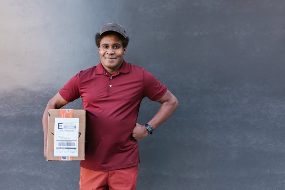 A smiling delivery person with medium brown skin and curly black hair, wearing a maroon polo shirt and matching red trousers, stands against a plain gray wall. They are holding a small cardboard parce