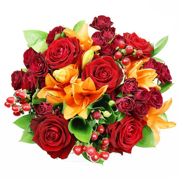 Captured from directly above, this sumptuous round bouquet sits against a spotless white field, revealing an artful, densely packed arrangement of deep red roses, vivid orange Asiatic lilies, and clustered burgundy spray roses. The large, fully bloomed red roses dominate the composition with their multilayered, velvety petals, each curl and shadow visible in the soft, even light. Bright orange lilies intersperse the reds, their trumpet silhouettes and visible stamens creating architectural movement while some blooms remain in the graceful promise of an unfurling bud. The smaller spray roses - concentrated yet delicate - introduce rich, wine-toned accents that increase the arrangement's depth and opulence. Scattered throughout are glossy hypericum berries whose small spherical forms catch and reflect light, offering jewel-like punctuation against the matte petals. Supporting foliage of wide, glossy ruscus or salal leaves peeks from beneath the flowers, its fresh green tones serving as a cooling backdrop and structural filler. The bouquet's overall impression is warm, celebratory, and tactile: the velvet of roses, the satiny lily petals, and the firm pop of berries together suggest both visual richness and a layered scent-spicy, citrus-tinged lilies tempered by the classic sweetness of roses. Pictured as a gift someone might carry to a celebration near Norton Road or to cheer a friend at a quiet walk in Nonsuch Park, this arrangement reads as an elegant declaration of affection, suited to anniversaries, autumn gatherings, or refined home décor.